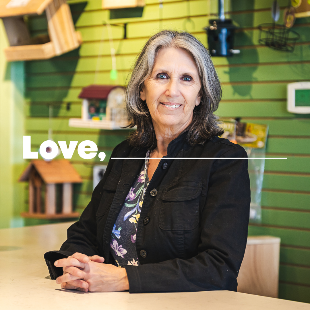 A smiling woman dressed in a black jacket, stands behind a counter in a shop with bird feeders on the wall behind her.